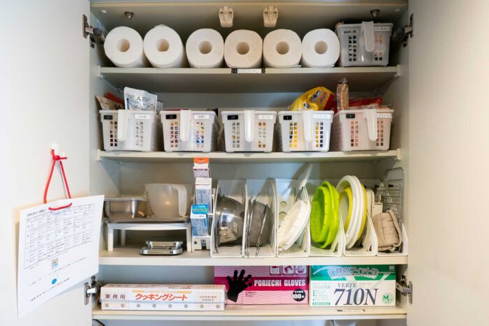 吊り戸棚の上段は、アイテム別に分けた食材を持ち手付きのボックスに入れて取り出しやすく。調理器具は片手でも取り出しやすいように収納。左の扉裏に「災害時の１週間献立表」を掛けている。